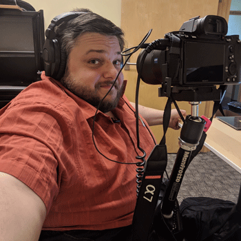 A white man in an orange shirt poses in headphones with a camera mounted to his chair.