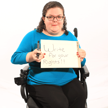 A white woman with curly brown hair wearing glasses, a teal sweater and black pants, sitting in a wheelchair and holding a marker and whiteboard on which is written, “Write for your rights!”